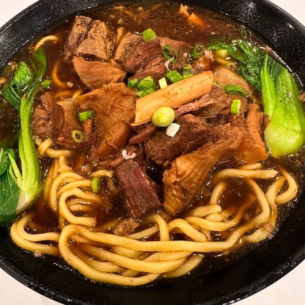 Close-up of Taiwanese beef noodle soup with bok choy, green onions, and thick noodles in a black bowl.